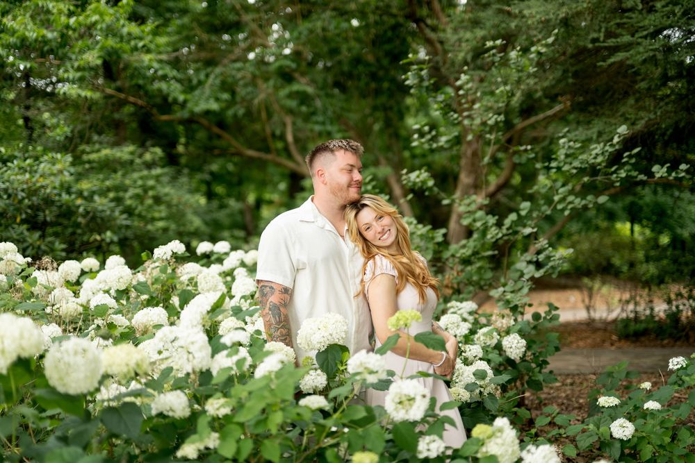 Couple embracing and smiling together among blooming white hydrangea bushes in a lush green garden.