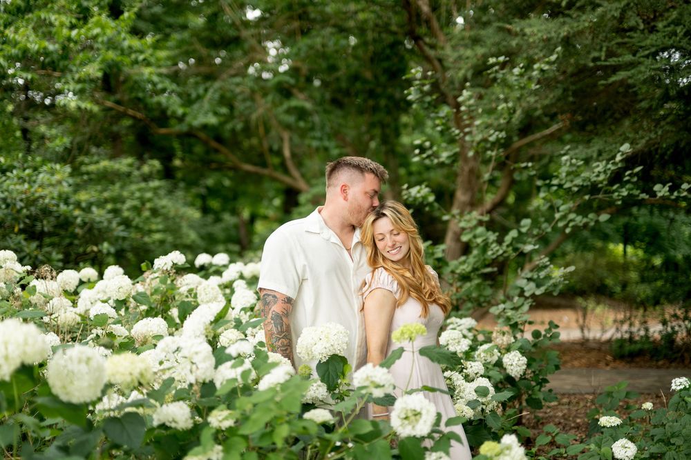 A couple shares a romantic moment surrounded by white hydrangea blooms in a lush green garden.
