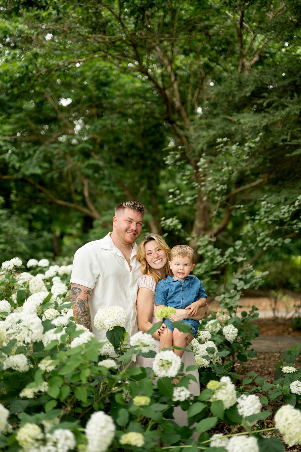 Happy family of three posing together among white hydrangea flowers in a beautiful green garden.
