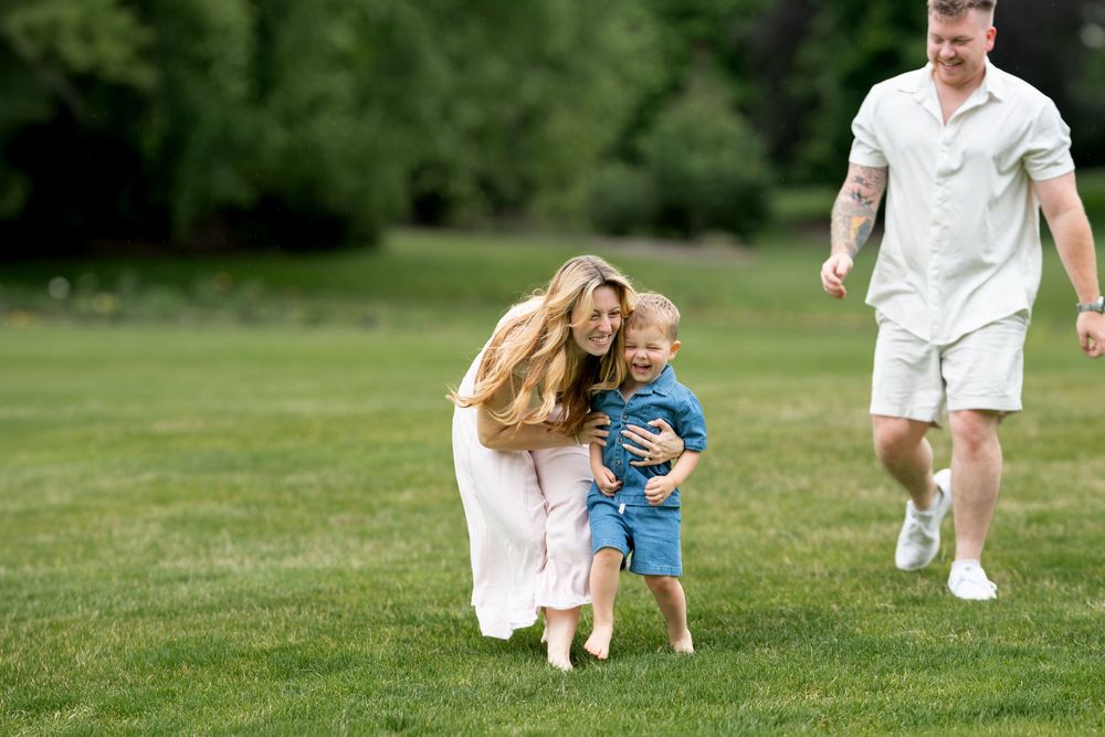 A laughing mother hugs a toddler boy on green grass as father jogs toward them outdoors.
