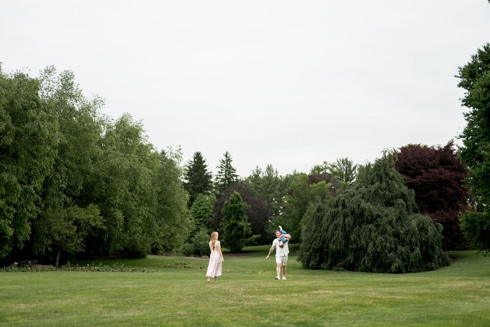 A couple stands apart on a vast green lawn surrounded by tall trees on an overcast day.