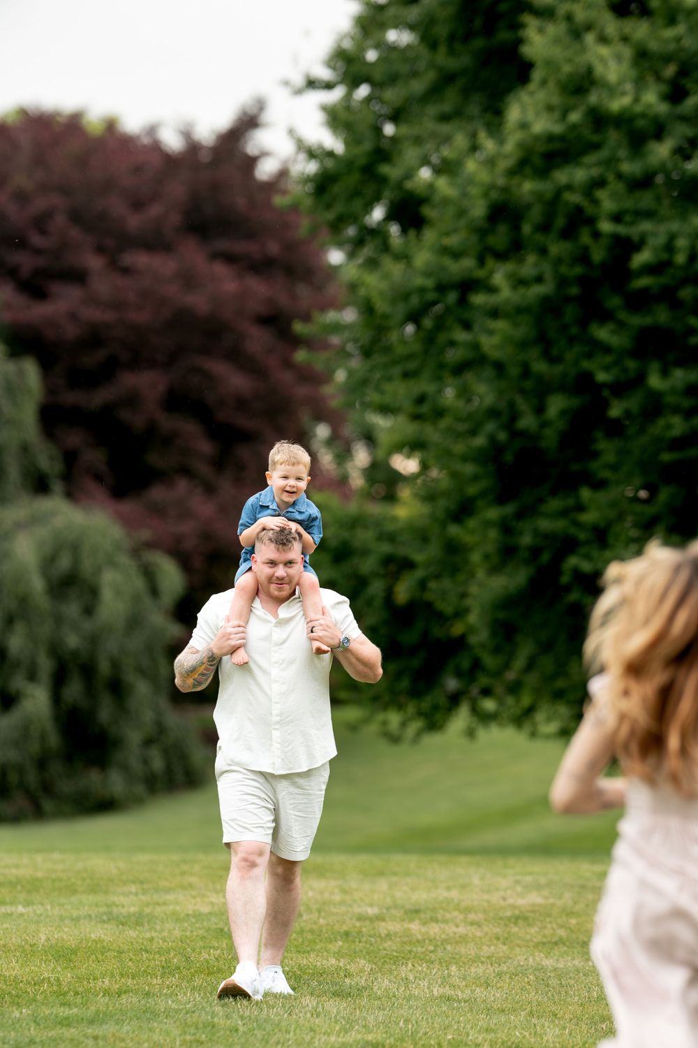 Smiling father carrying young son on shoulders while walking toward mother in a green park.