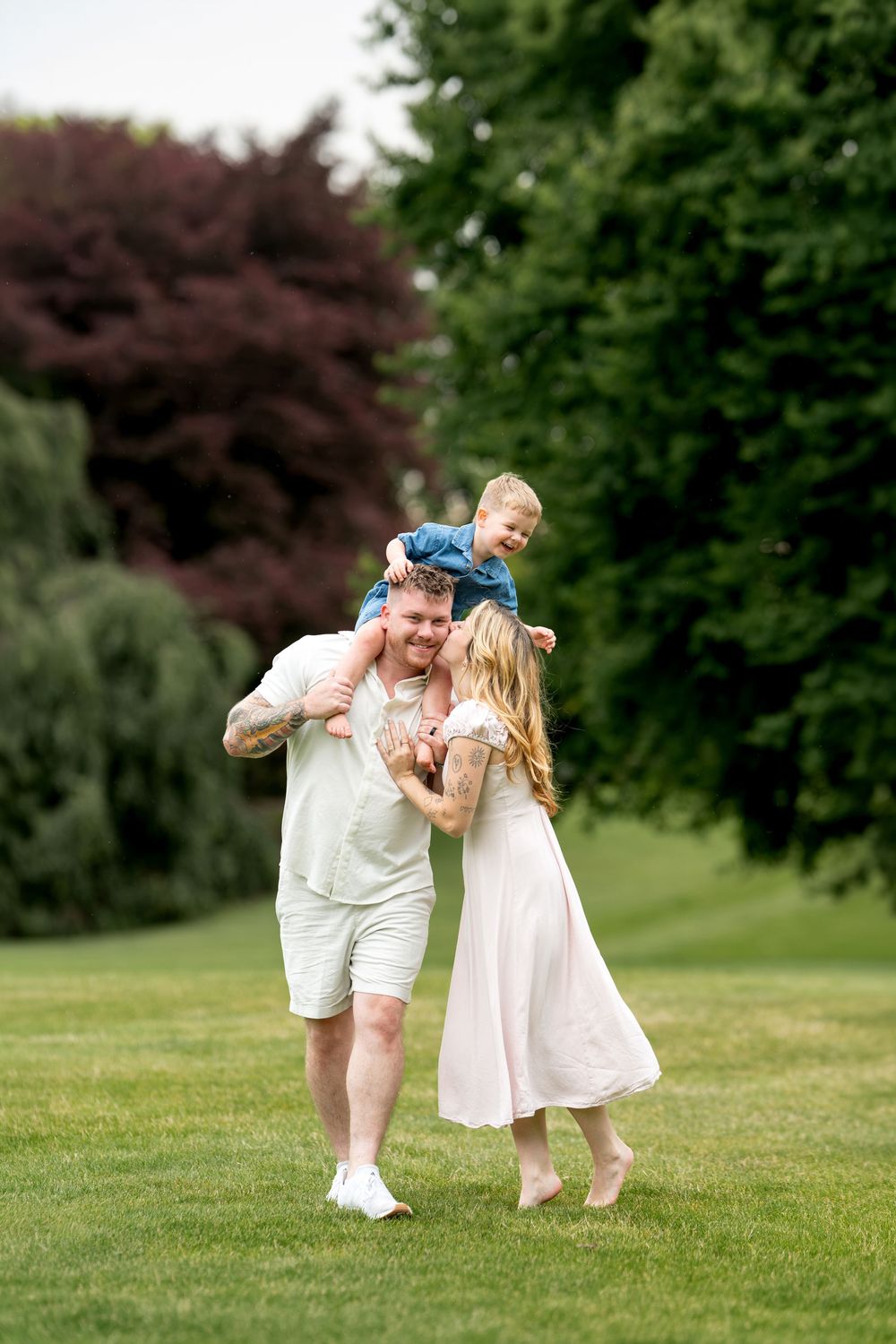 Joyful family with tattooed father carrying toddler on shoulders while mother walks beside them on green lawn.