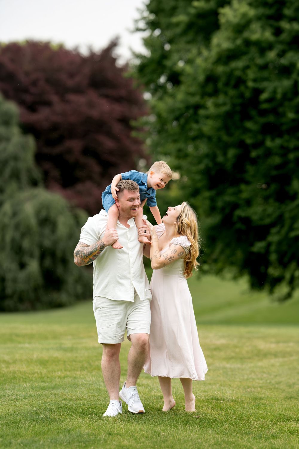 A happy family laughs together as father lifts toddler on shoulders with mother beside them outdoors.
