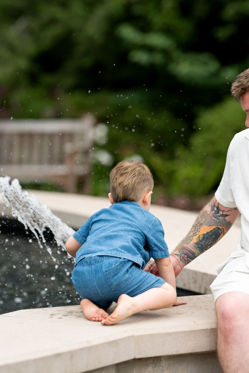 Curious toddler reaching toward a garden fountain while tattooed father watches closely nearby.