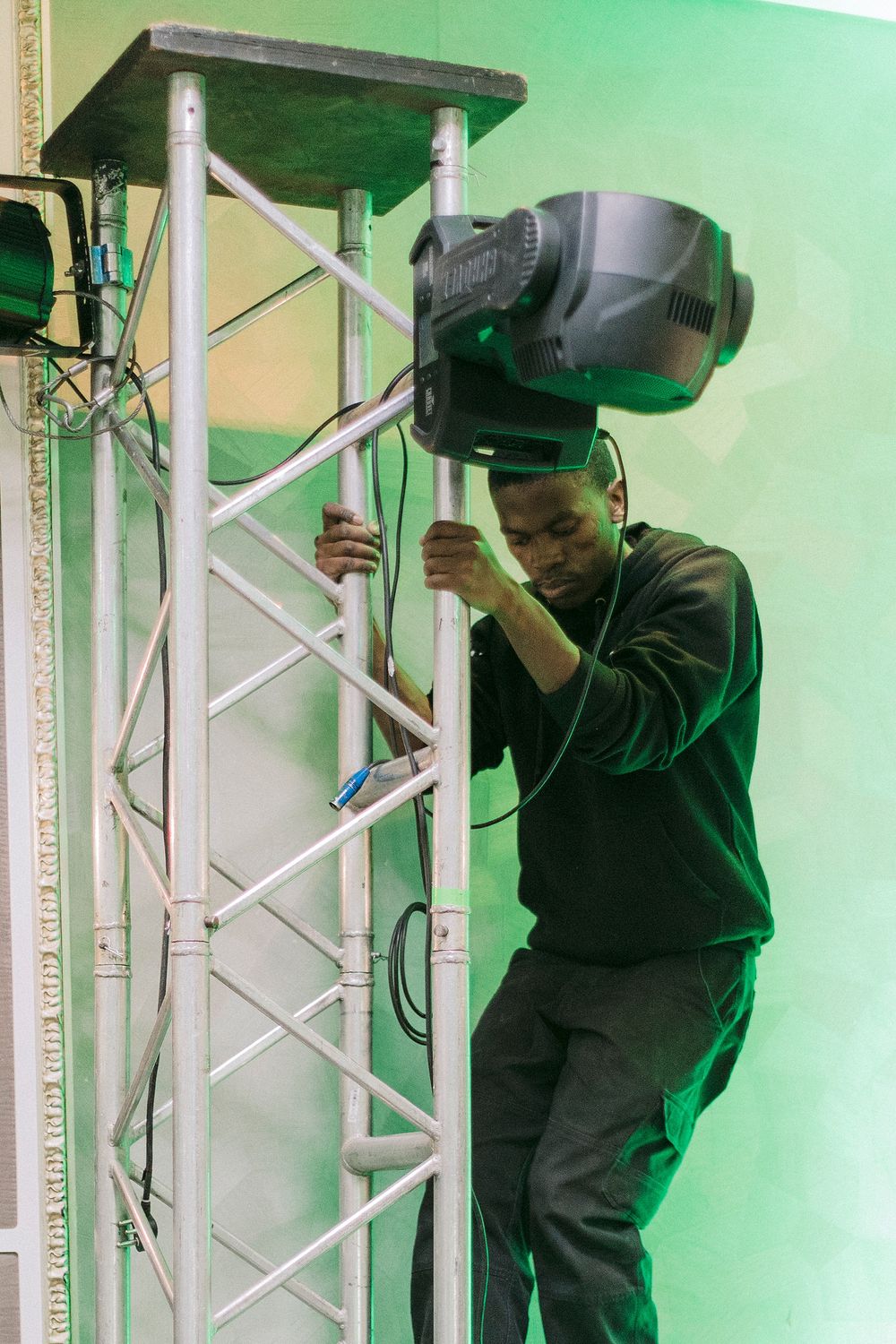 A technician works on a moving head spotlight mounted on a silver truss in a green-lit studio.