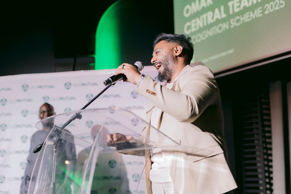 A man in a beige suit speaks energetically into a microphone at a corporate awards recognition event.
