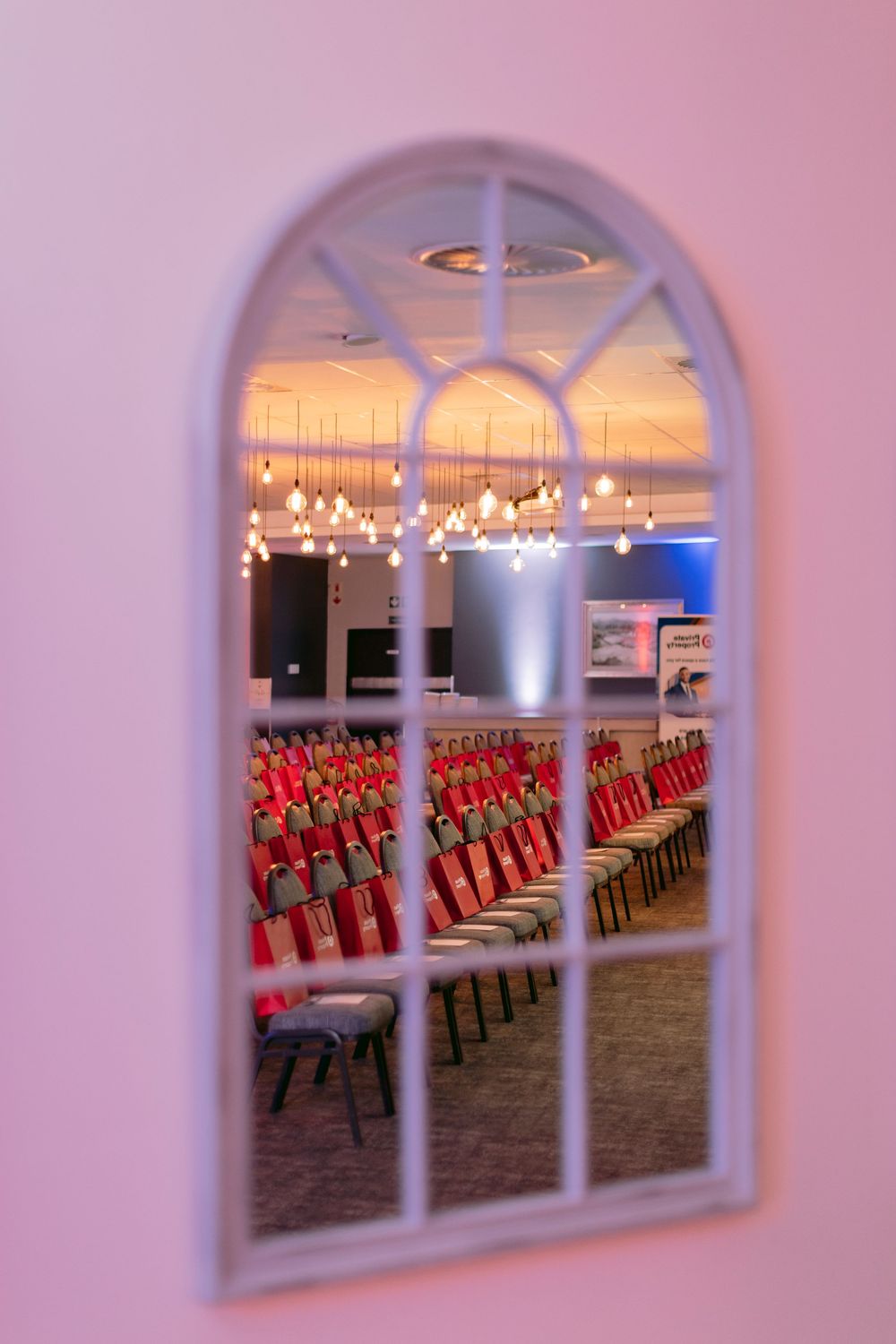 Arched window mirror reflecting rows of red chairs and warm Edison bulb lighting in an elegant event venue.