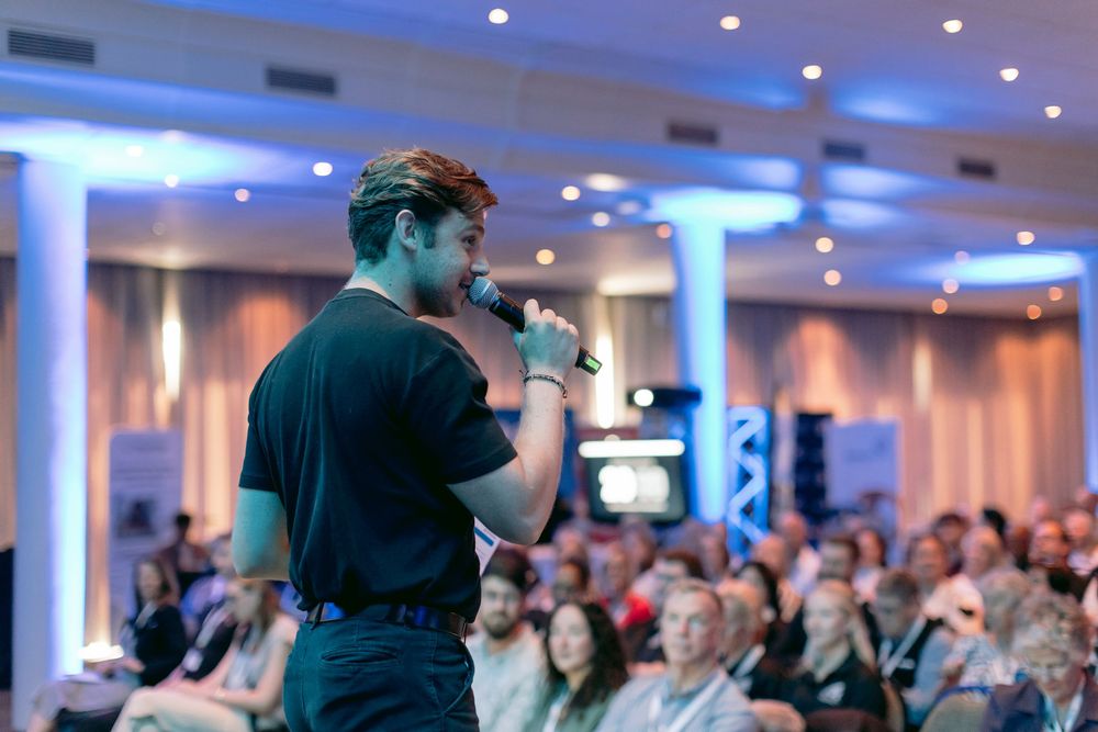 Male speaker holding microphone addressing a large seated audience in a blue-lit event hall.