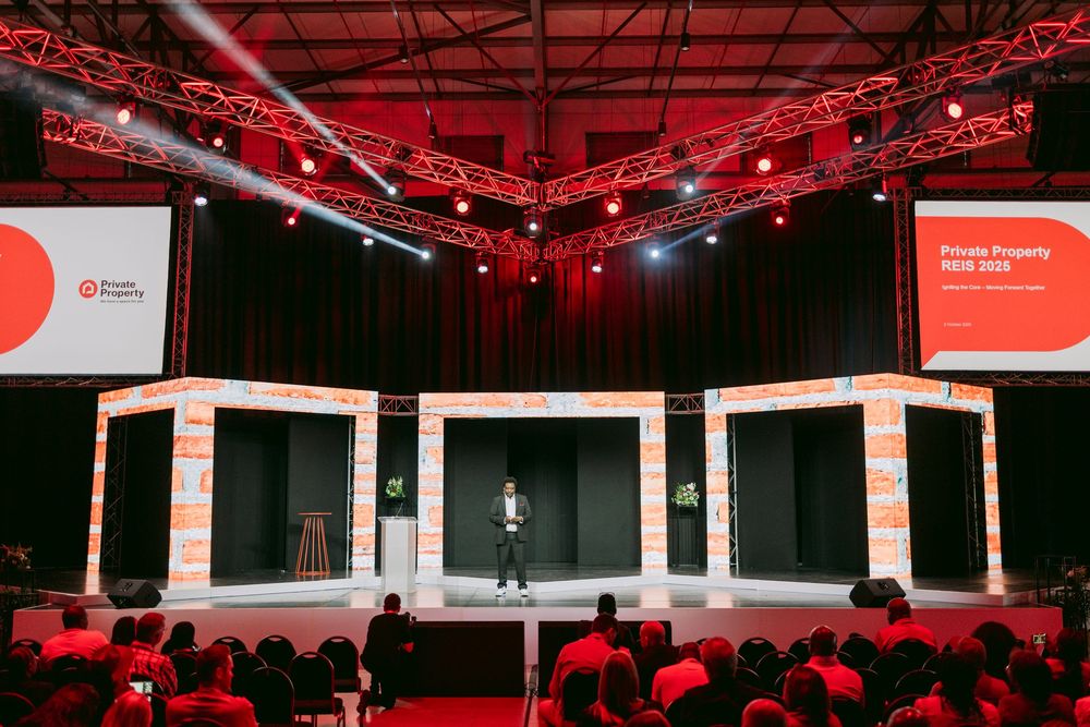 A speaker stands on a large red-lit conference stage with bold branding and an audience seated in the foreground.