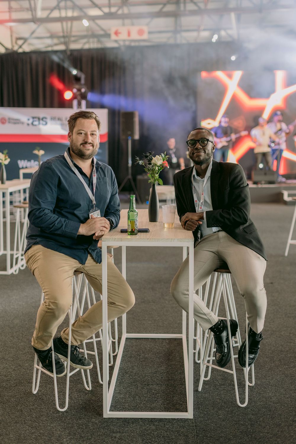 Two men sitting at a high white table at a corporate event venue with stage lighting.