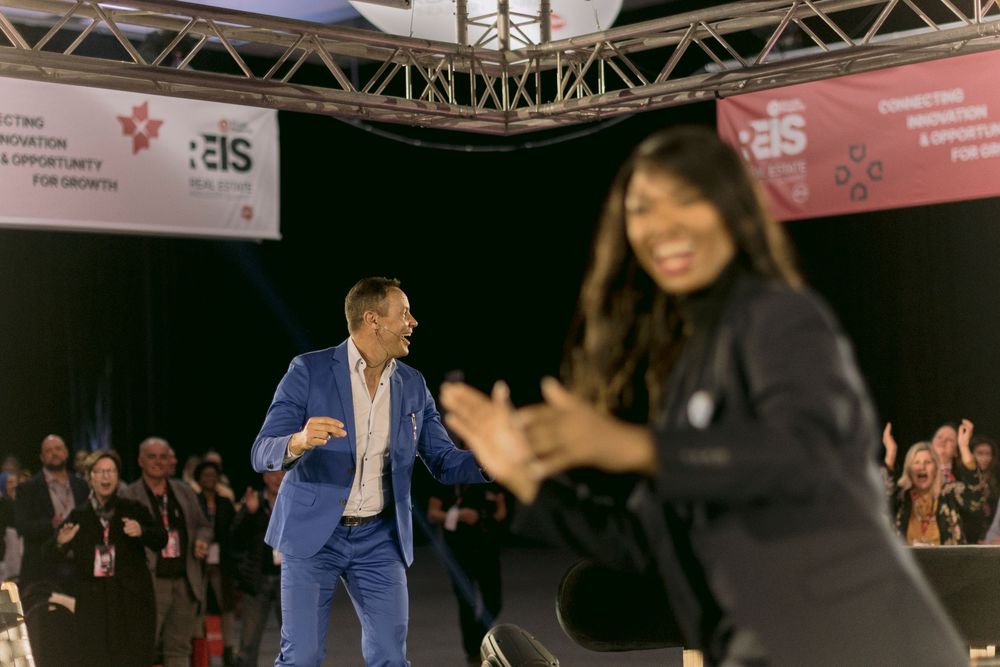 Man in blue suit dancing on stage with a woman clapping in the foreground at a lively conference event.