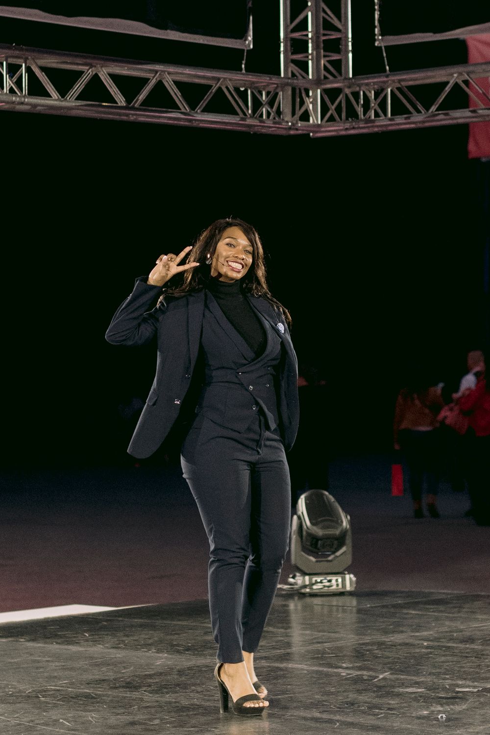 Woman in navy blue suit making peace sign on stage at conference event.