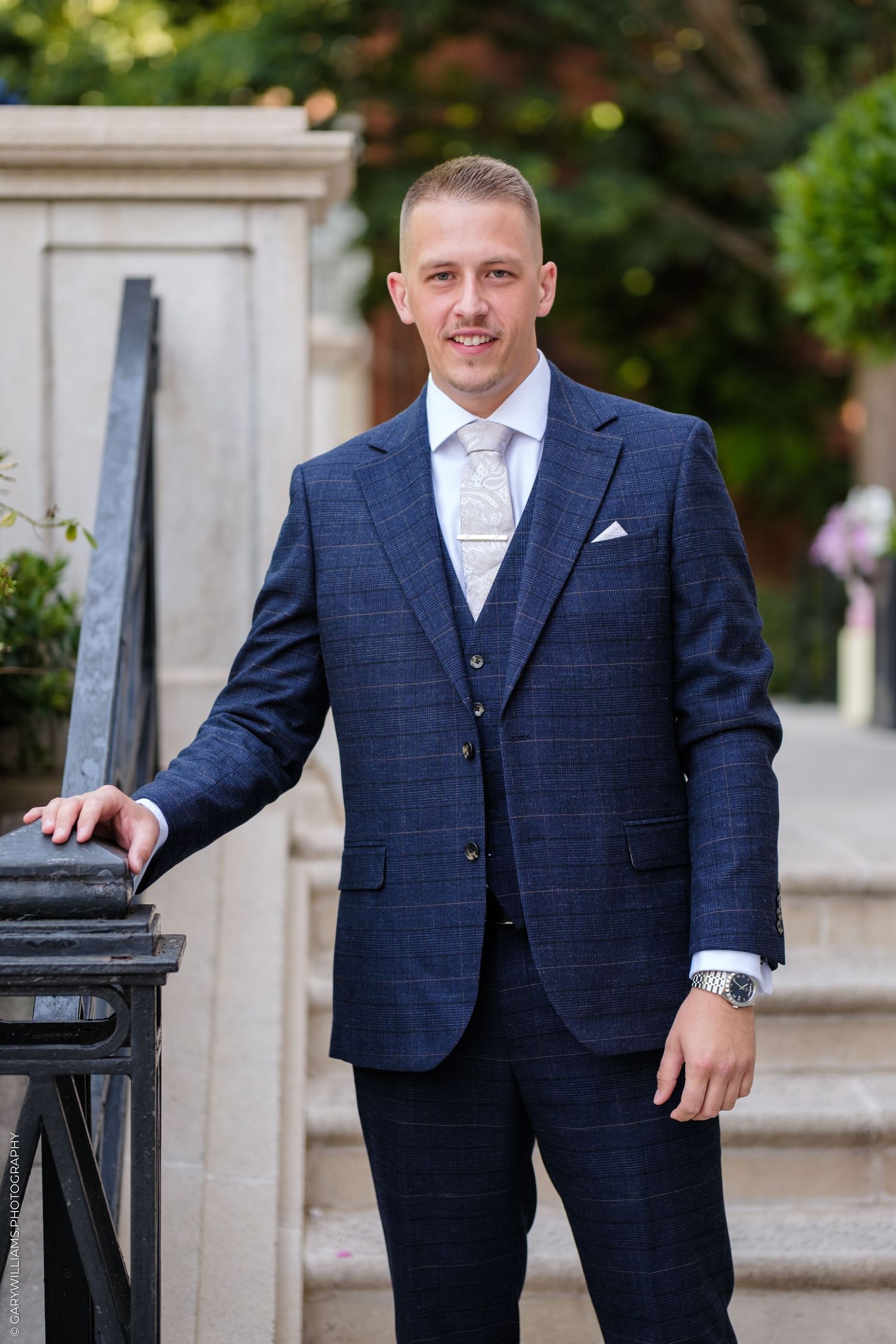 A smiling man in a navy blue three-piece suit with a light tie poses on stone steps outdoors.