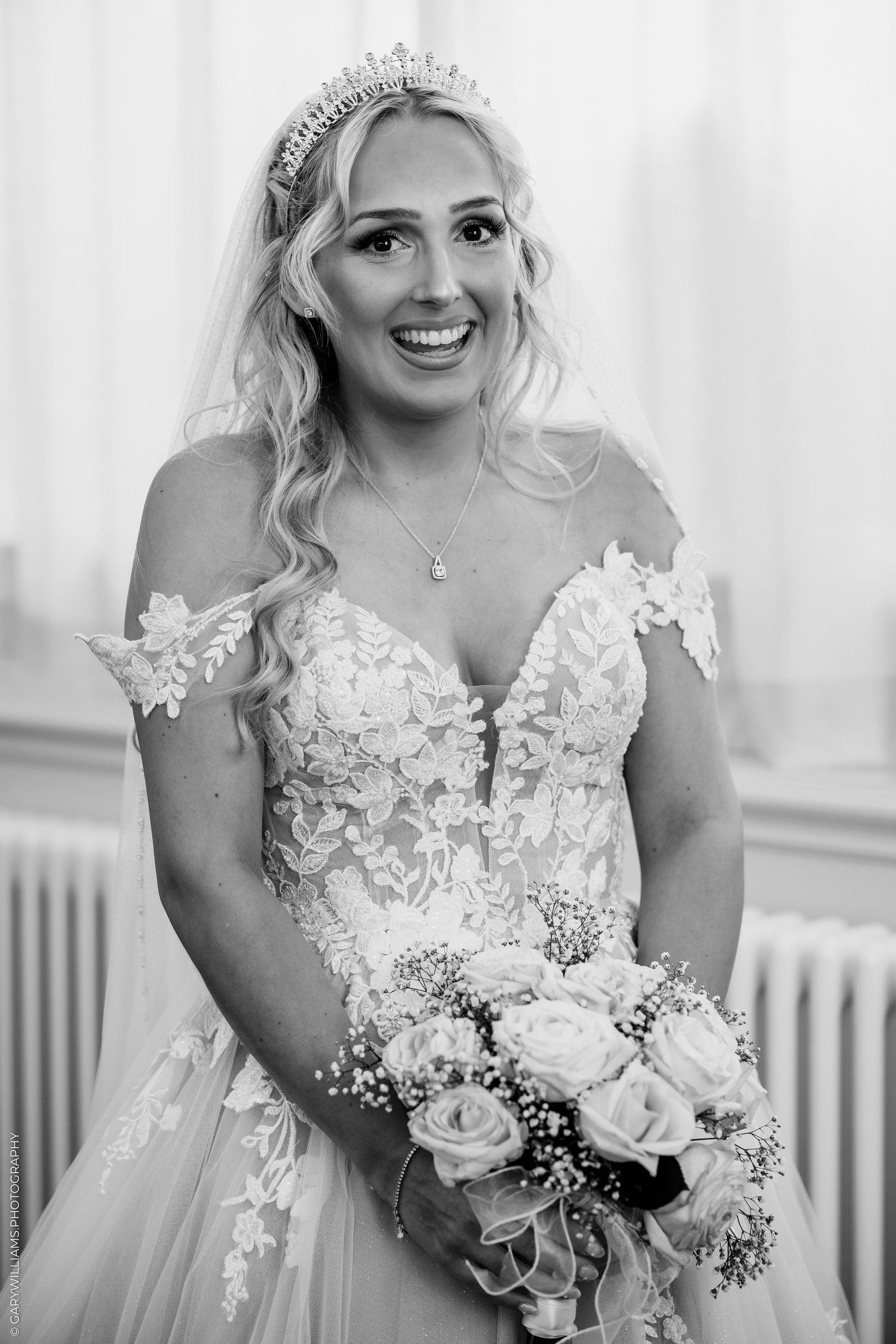 Smiling bride in lace off-shoulder gown and tiara holds rose bouquet in elegant black and white wedding portrait.