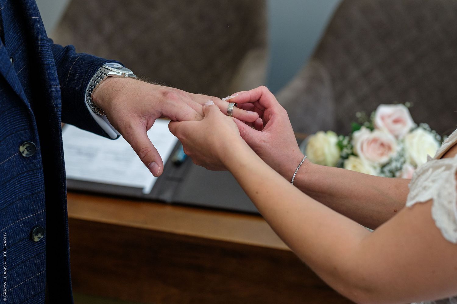 Bride placing a wedding ring on groom's finger during a wedding ceremony, with a bouquet of roses in the background.