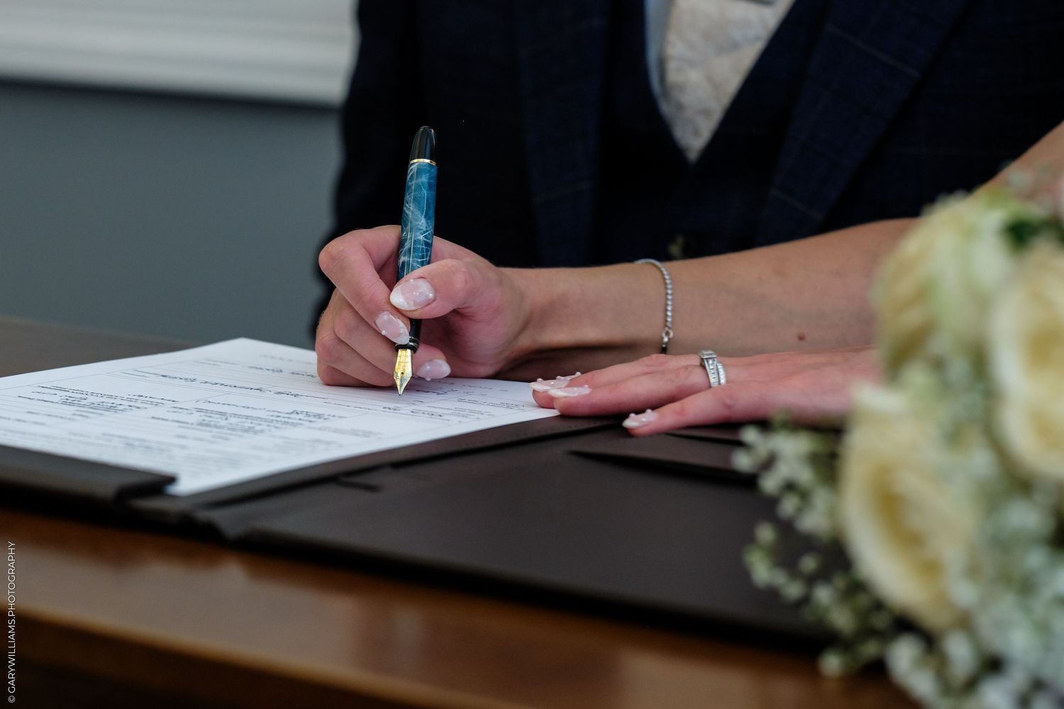 Bride signing wedding certificate with a blue pen, wearing a diamond ring and bracelet, with a bouquet nearby.