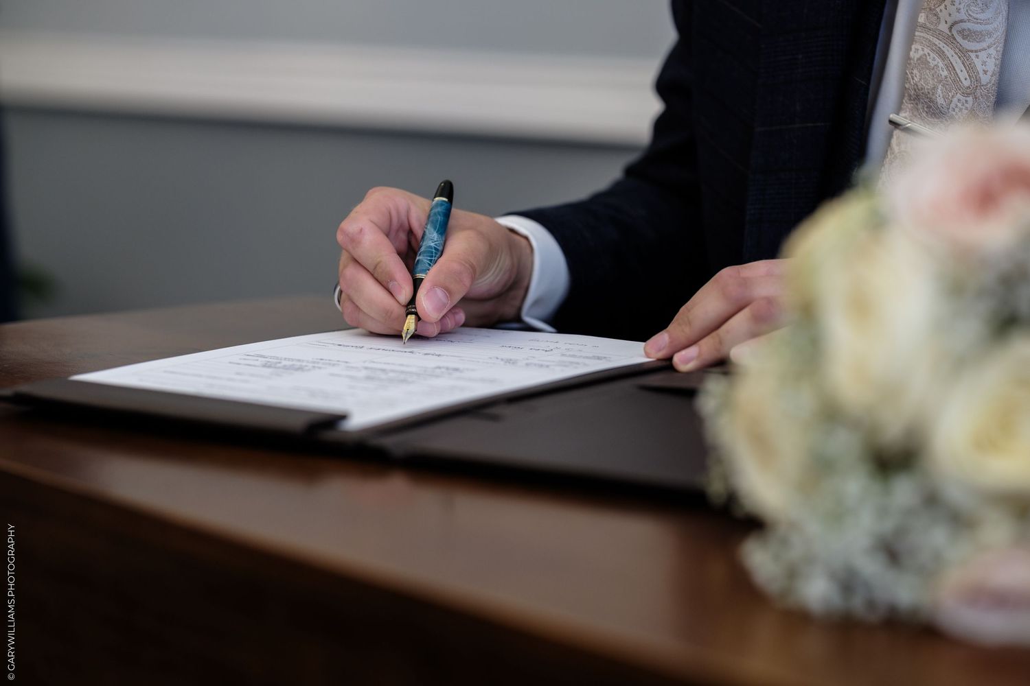 Person in suit signing wedding certificate with elegant pen, floral bouquet in foreground on wooden table.
