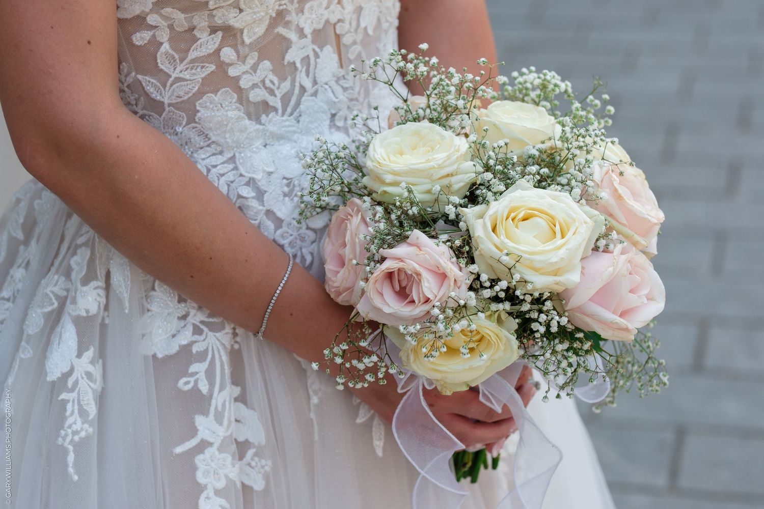 Bride holding a delicate wedding bouquet of white and pink roses with baby's breath, wearing a lace bridal gown.