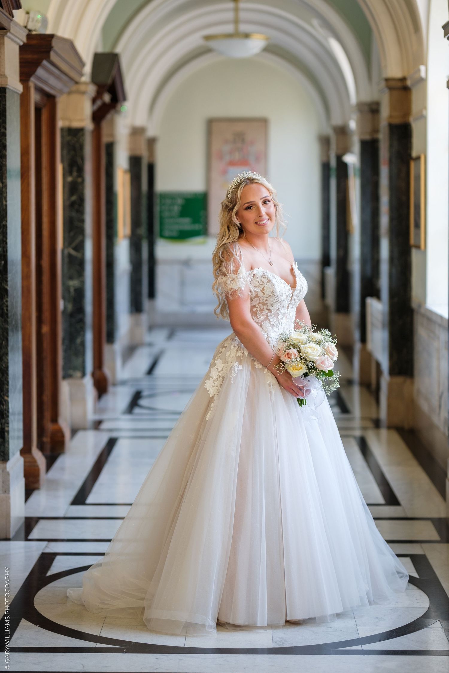 Bride in a white ball gown holding a bouquet poses in an elegant marble hallway with tall columns.