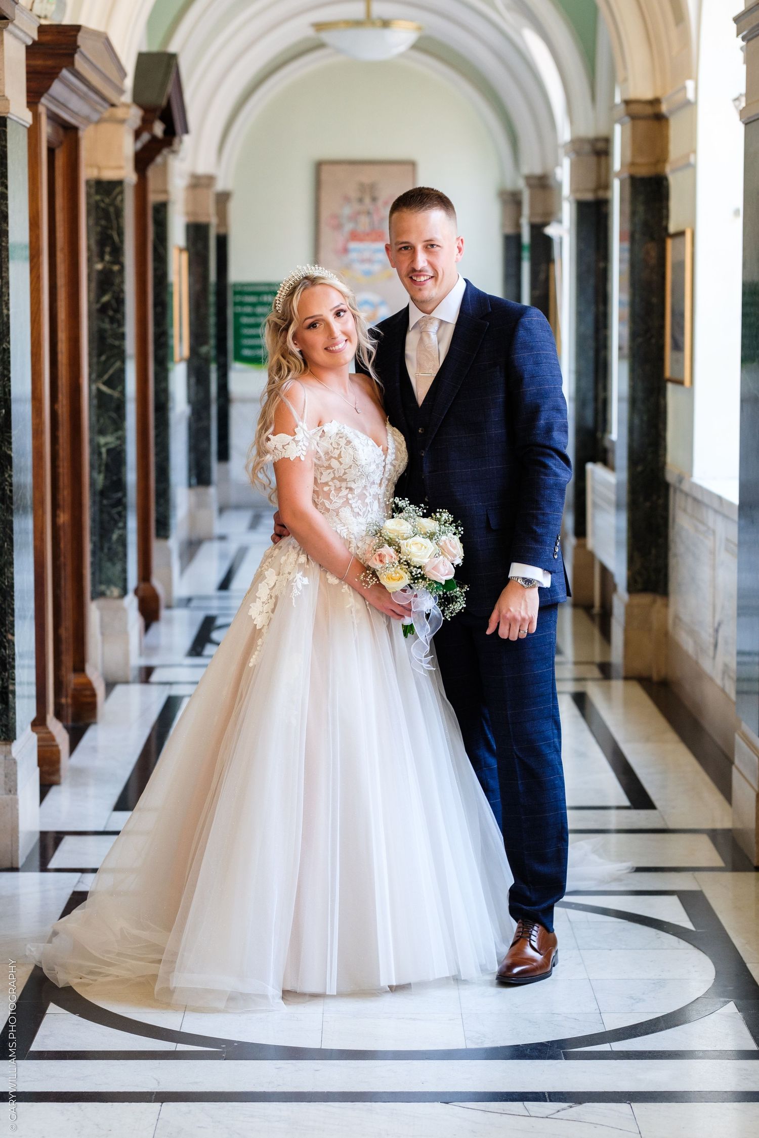 Bride in white gown holding bouquet stands with groom in navy suit in elegant marble hallway.