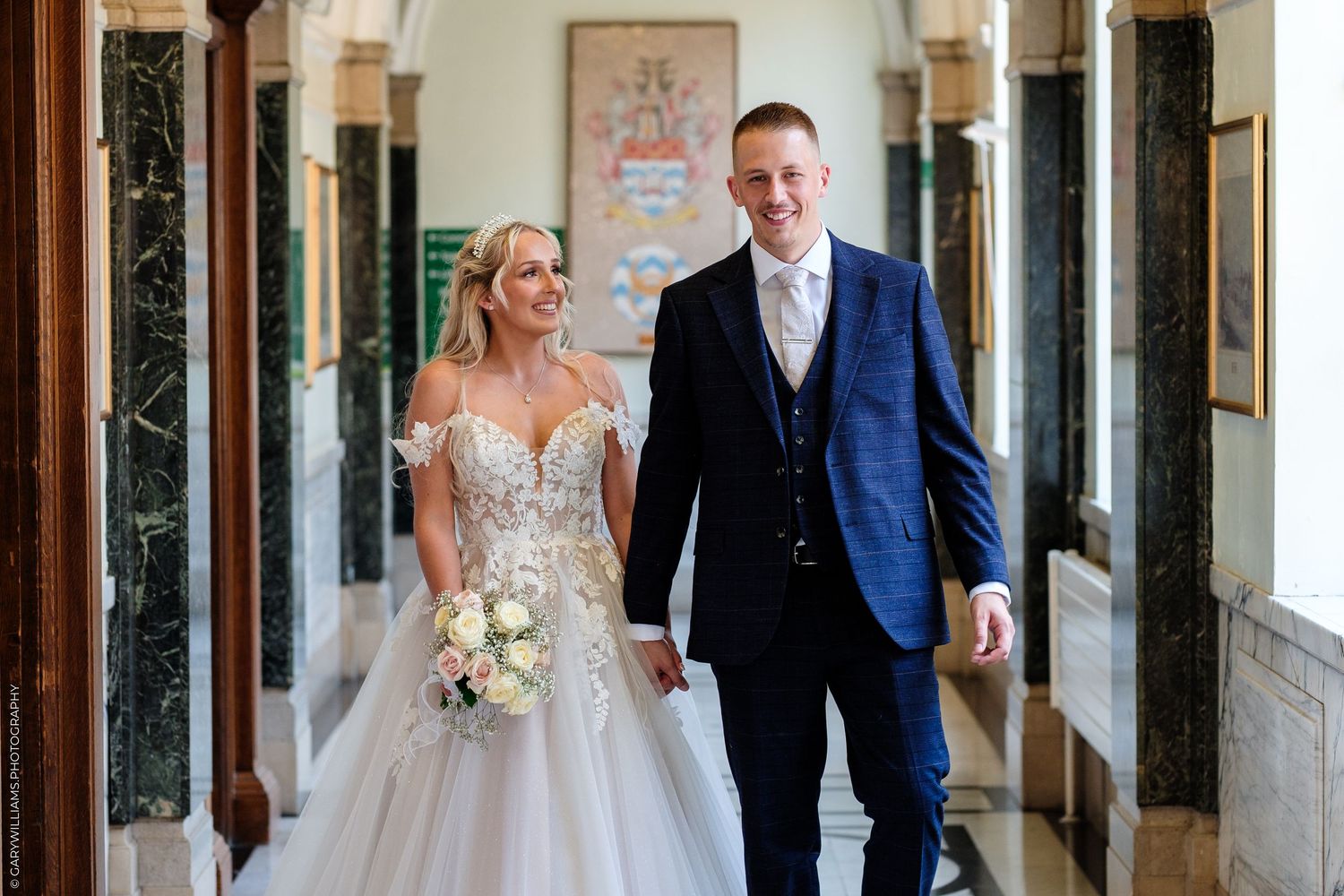 Bride and groom walking hand in hand down an elegant hallway, bride in lace gown holding bouquet.
