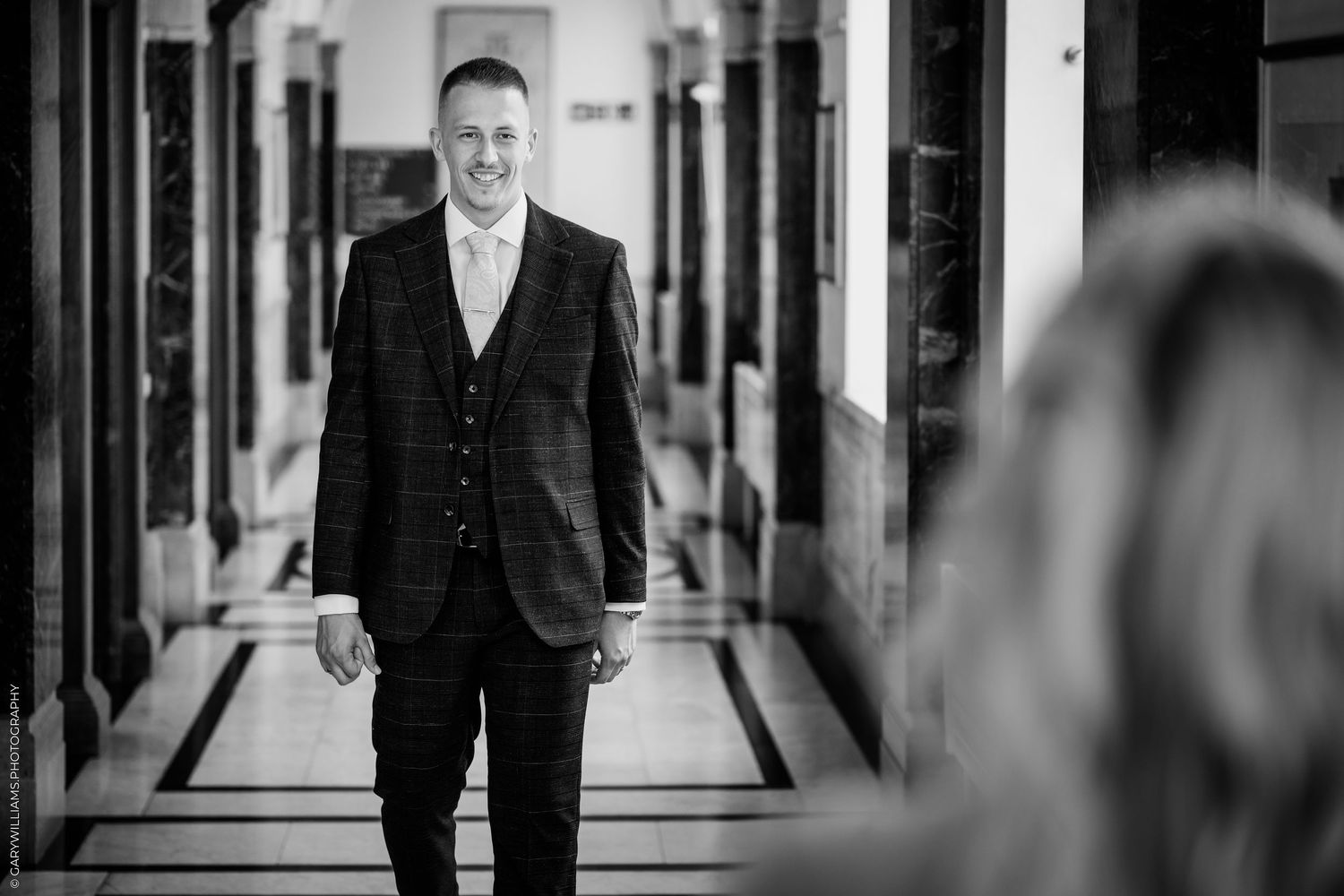 Smiling groom in a plaid suit walks down an elegant hallway in a black and white wedding photo.
