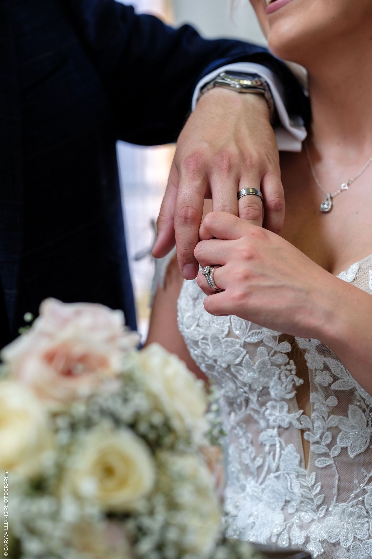Bride and groom holding hands displaying wedding rings, with a white lace bridal gown and bouquet of roses visible.
