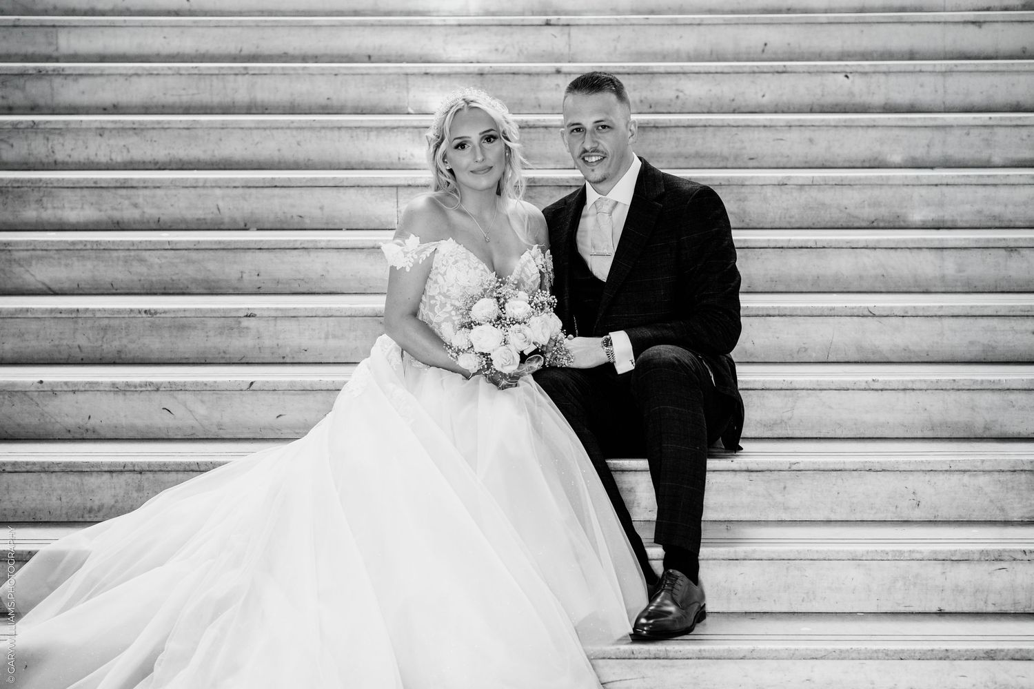 Black and white photo of a bride and groom sitting on marble steps, bride holding a bouquet in a ball gown.