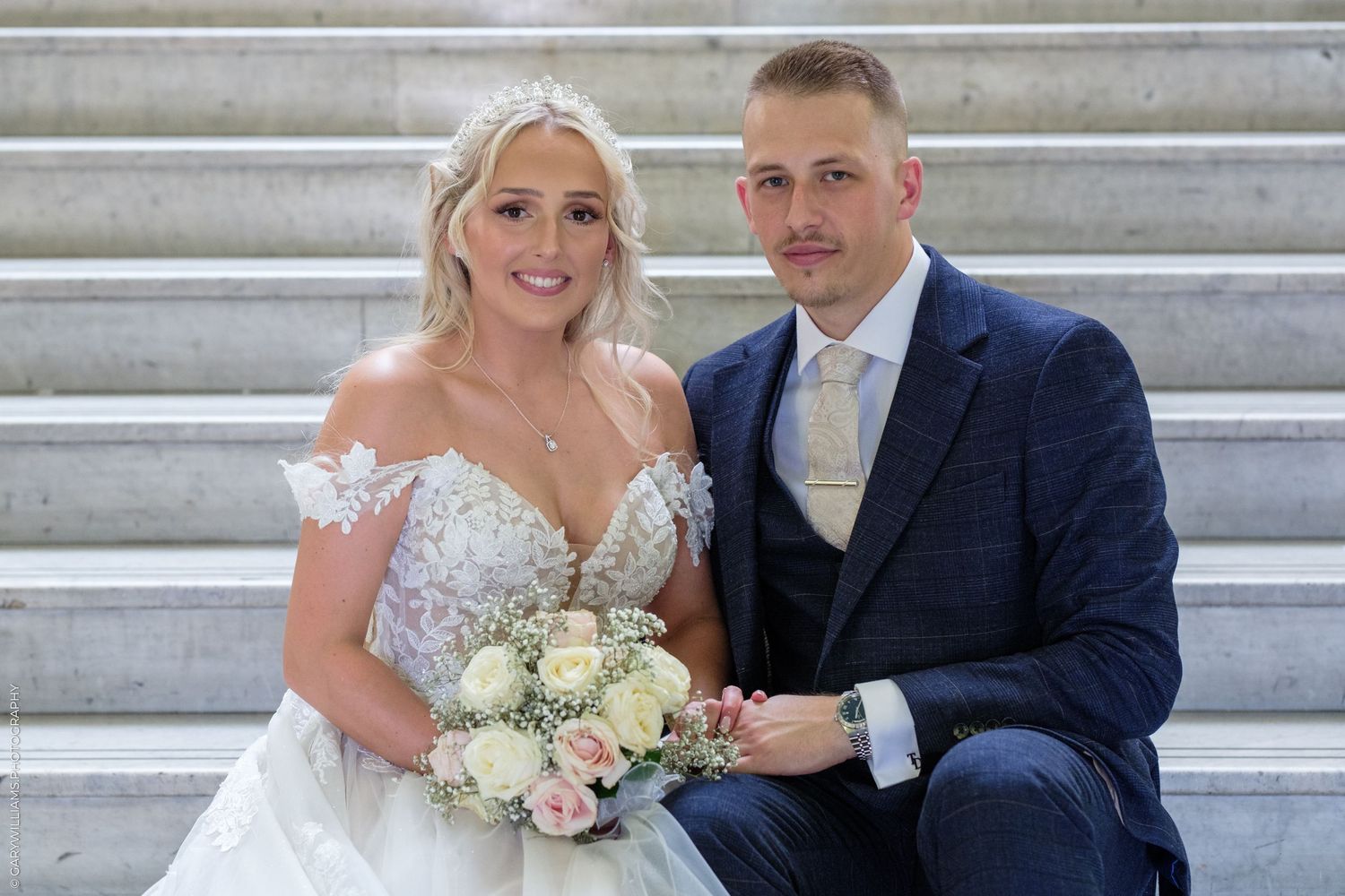 Bride and groom posing on marble steps, bride in lace gown holding pink bouquet, groom in navy suit.