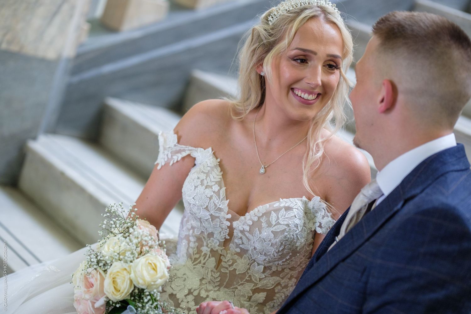 Bride in lace off-shoulder gown holding roses bouquet smiles at groom in blue suit on elegant staircase.