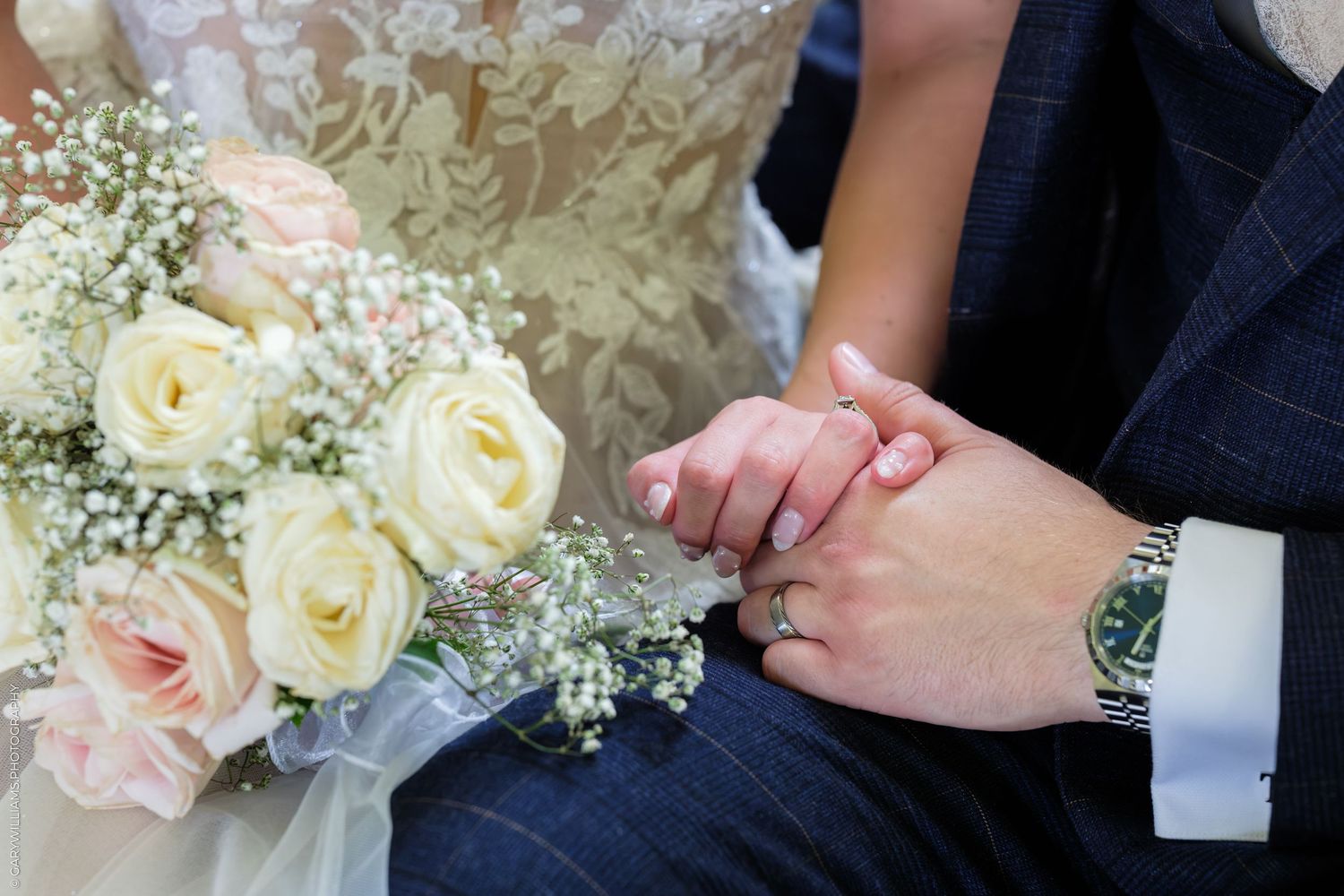 Bride and groom holding hands with wedding bouquet of white roses and baby's breath, groom wearing a watch.