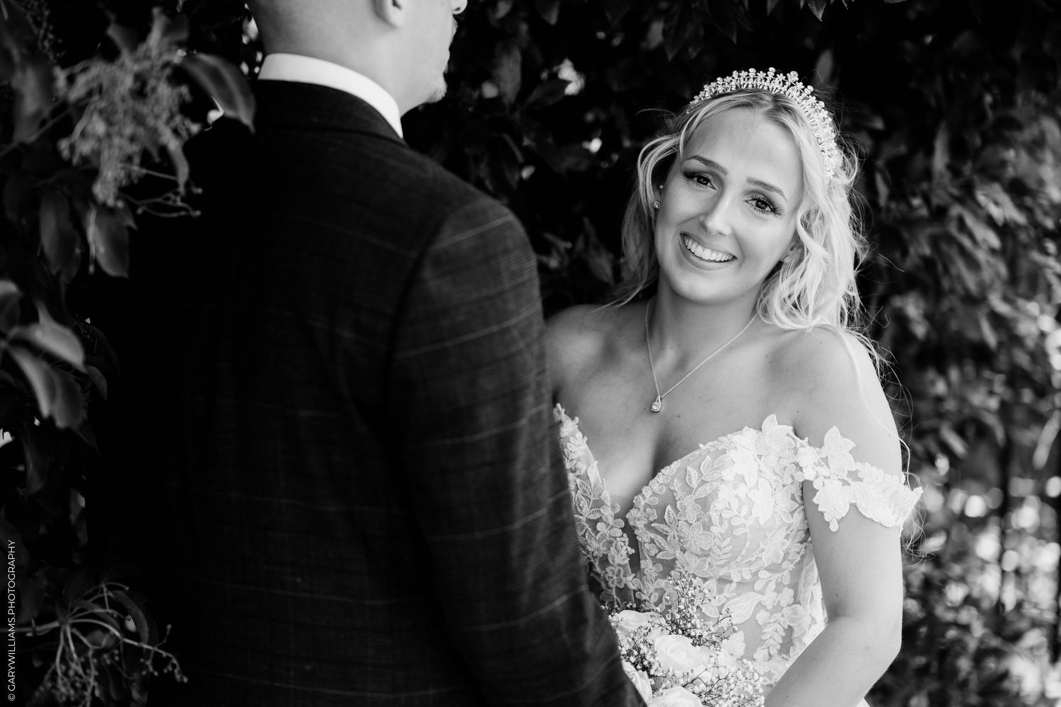 Black and white photo of a smiling bride in lace gown and tiara facing groom outdoors at wedding ceremony.