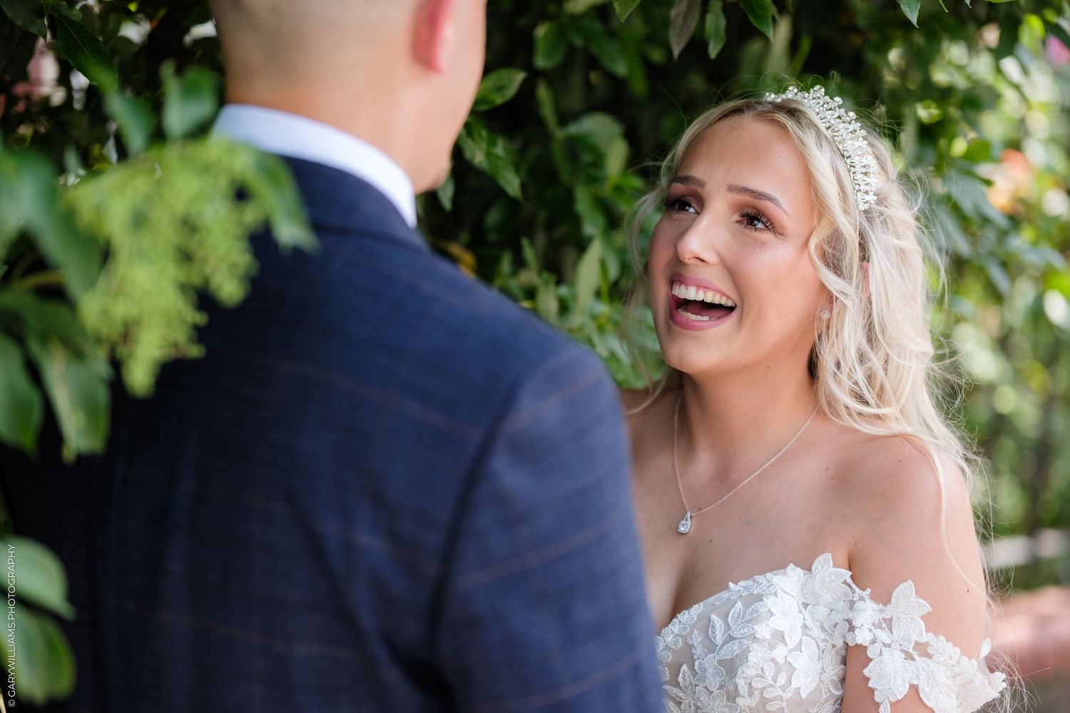 Joyful bride in lace gown laughing during outdoor wedding ceremony surrounded by lush green foliage.