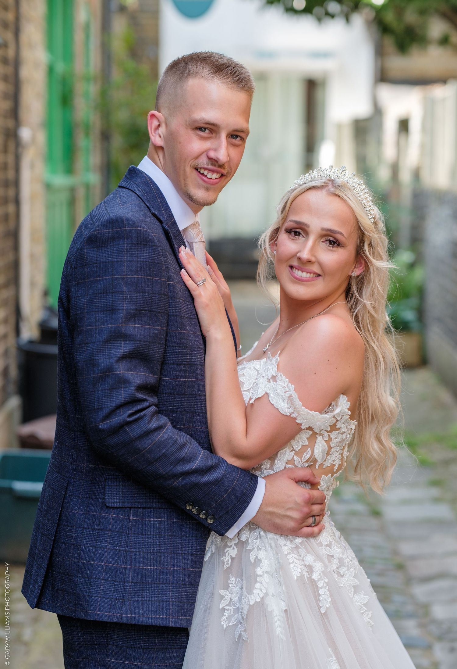A smiling bride in a lace wedding dress and groom in a navy suit pose together outdoors on their wedding day.
