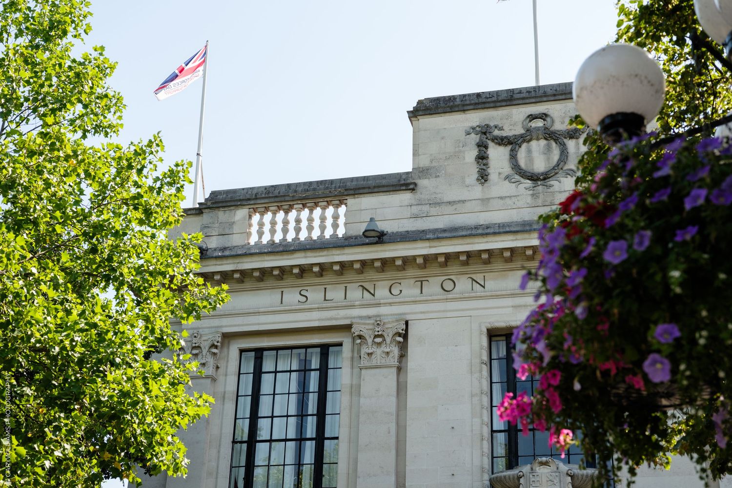 Islington Town Hall building with Union Jack flag, surrounded by purple flowers and green trees on a sunny day.