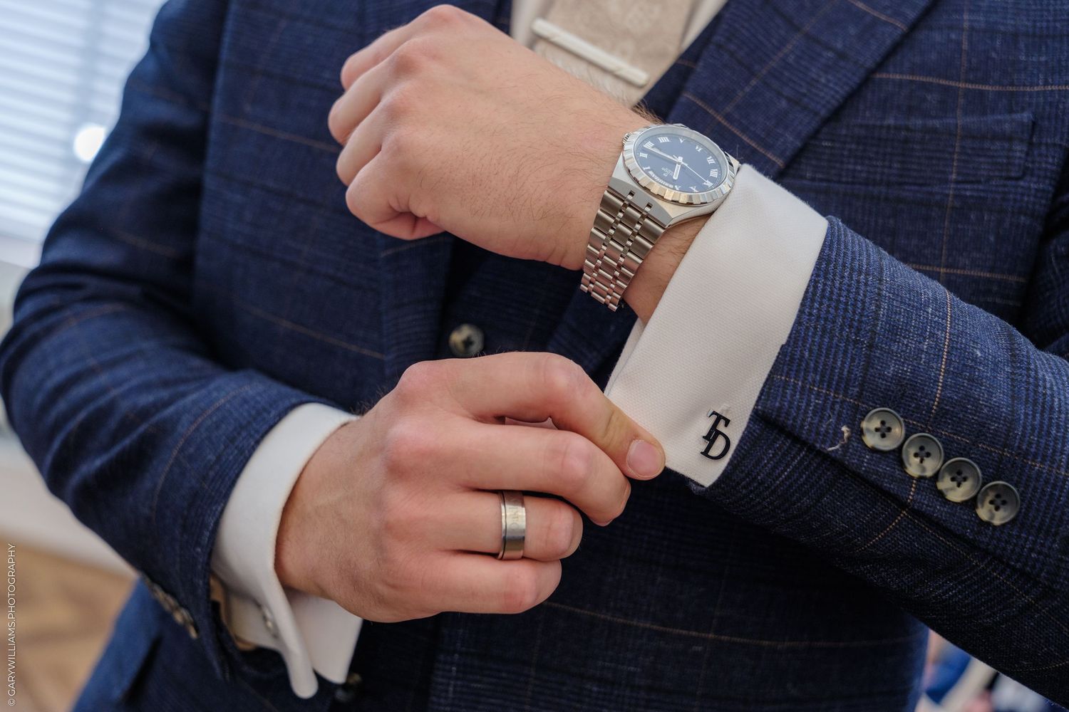 Man in blue plaid suit adjusting cufflinks, wearing a silver Rolex watch and ring on his finger.