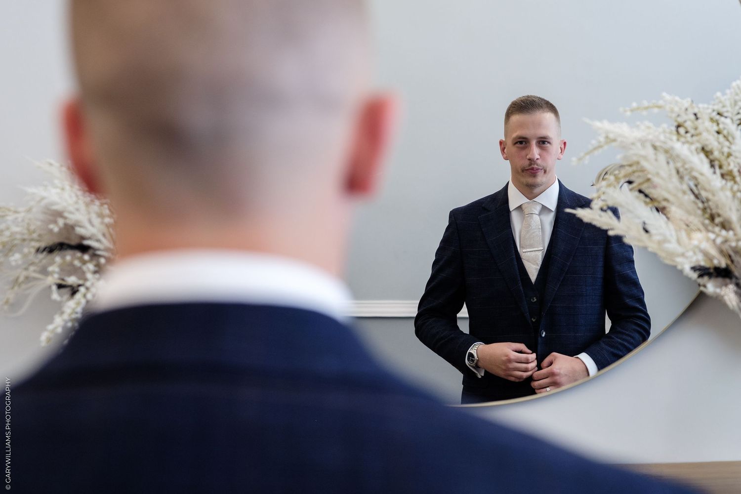 Groom in navy suit adjusting jacket while checking his reflection in an oval mirror on wedding day.
