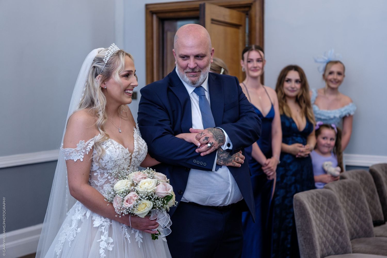 Bride in lace gown holding white bouquet stands with suited man, guests in blue dresses visible behind at wedding ceremony.
