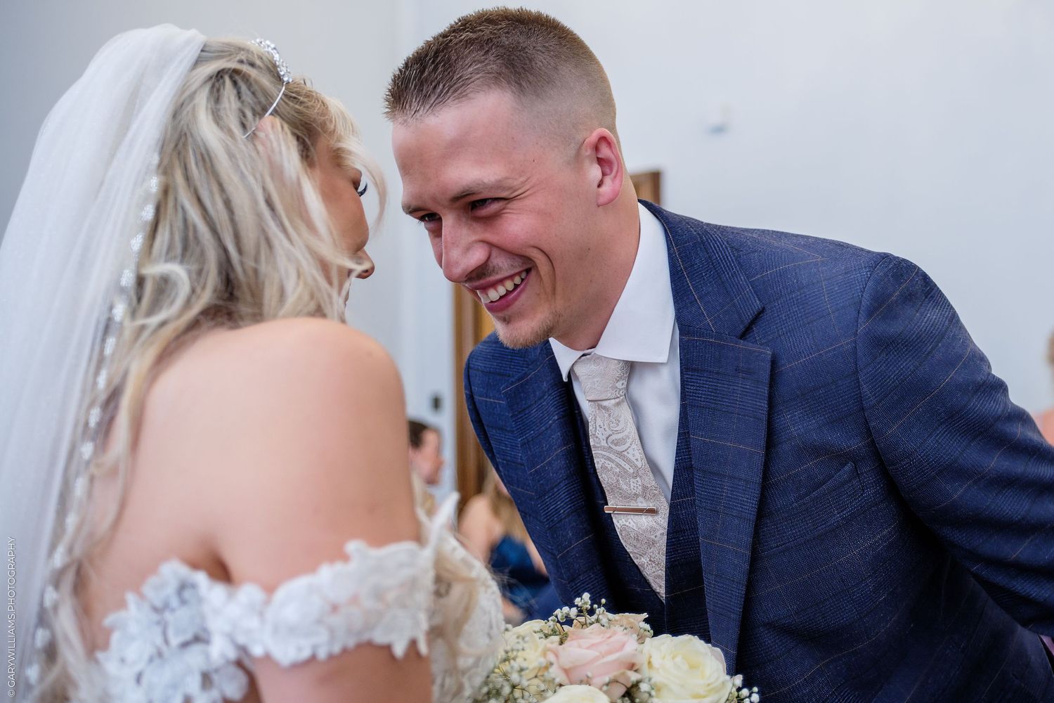 A joyful groom in a blue suit laughs with his bride in a white lace dress during their wedding ceremony.