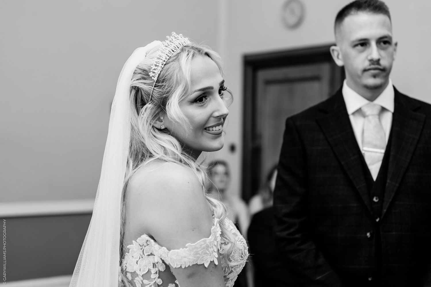 Smiling bride in lace gown and tiara veil stands with groom during wedding ceremony.