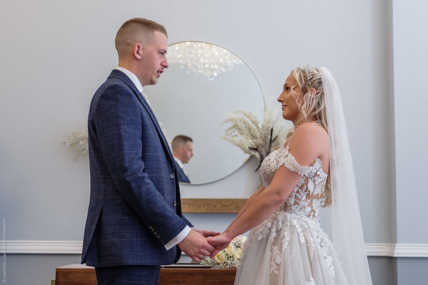 Bride and groom holding hands and smiling at each other during wedding ceremony in elegant venue.