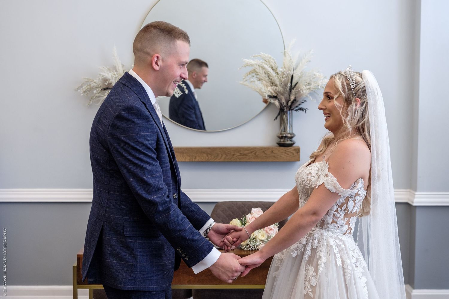 A bride and groom hold hands during their wedding ceremony, with the bride in a lace gown and groom in a navy suit.