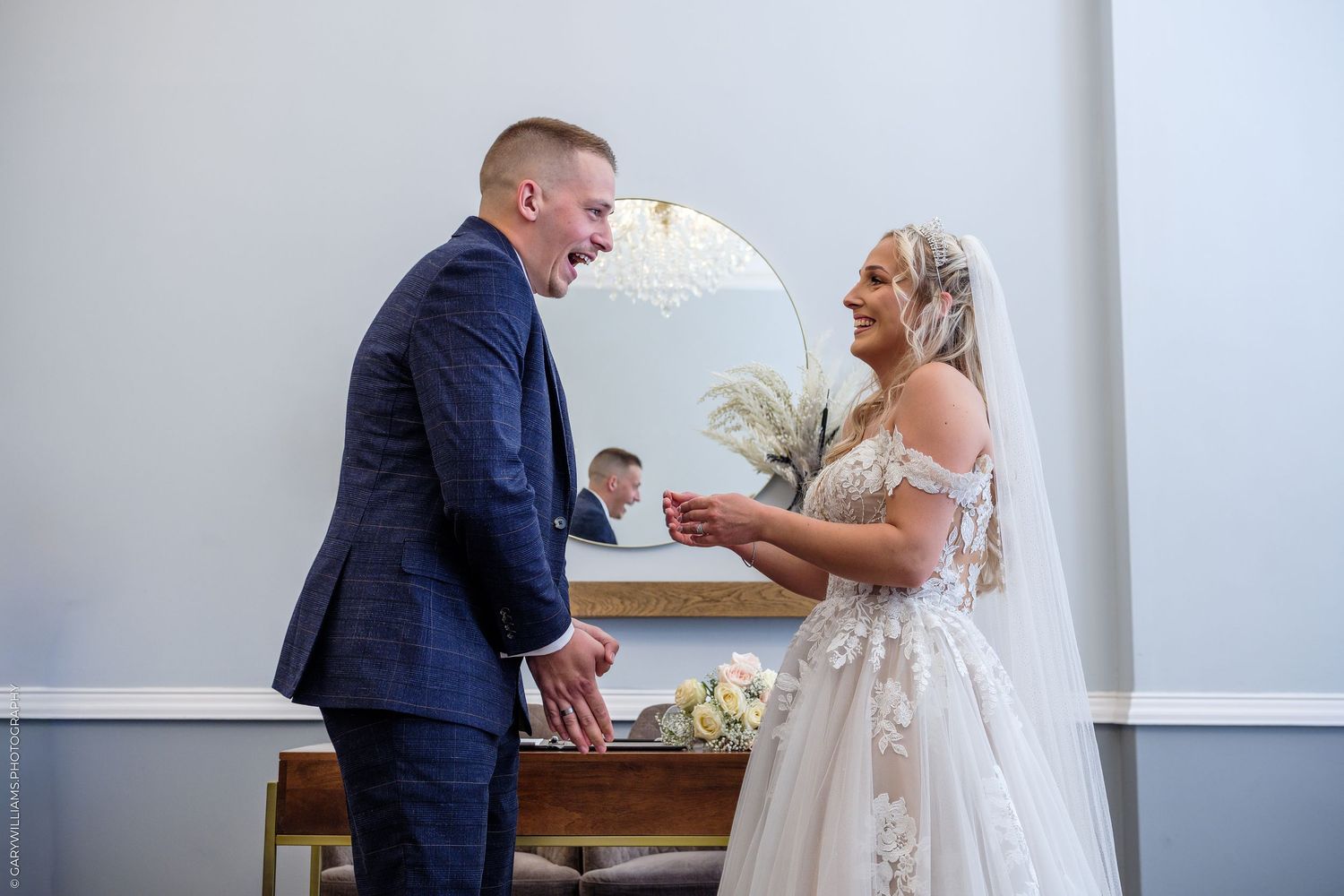 Bride and groom share a joyful moment together indoors, with the bride in a lace gown and groom in a navy suit.