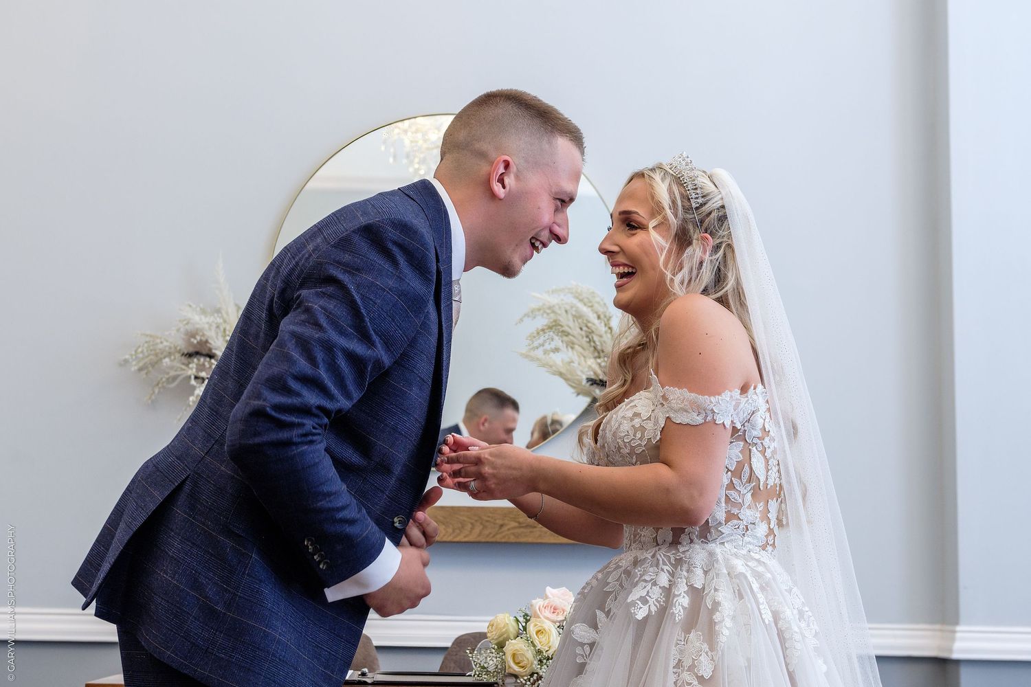 Bride and groom share a joyful laugh during their wedding ceremony in an elegant white venue.