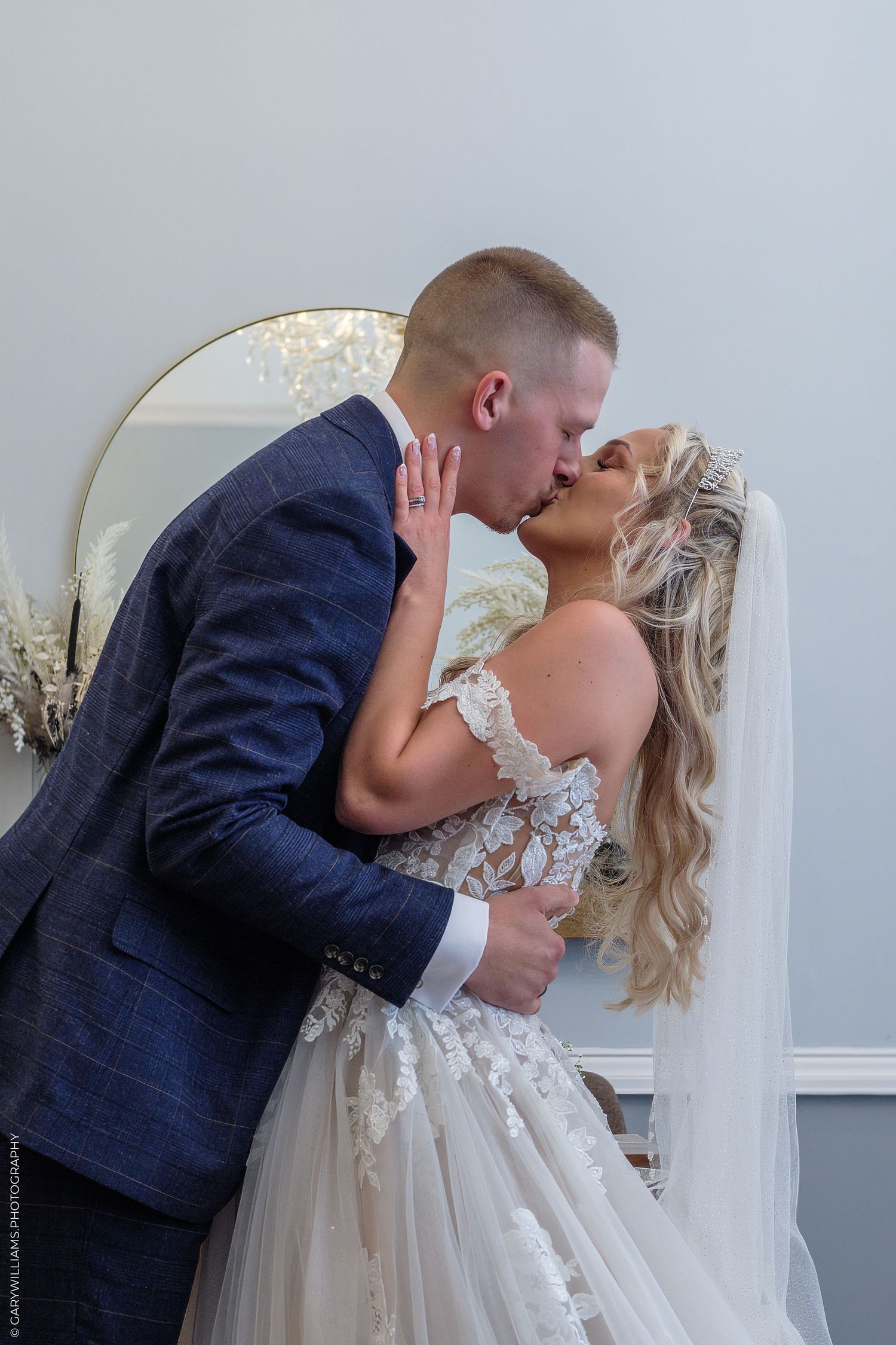 A bride and groom share a romantic kiss in their wedding attire, with a gold mirror and soft décor in the background.