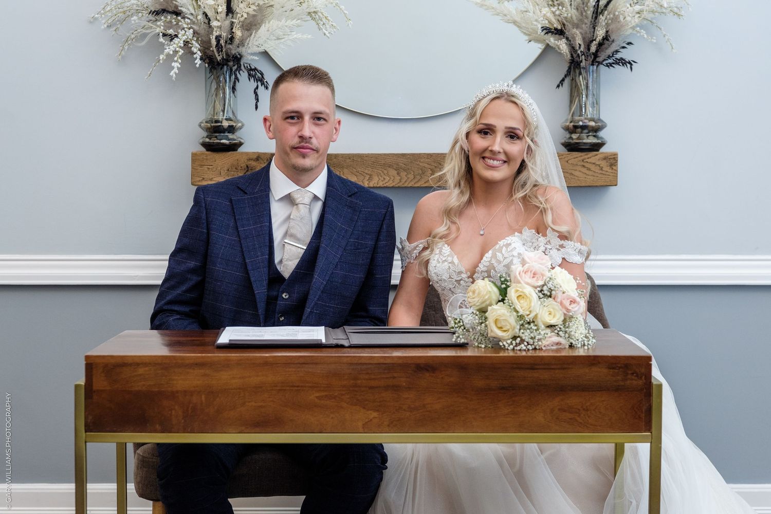 Bride and groom seated at wooden signing table, bride holding bouquet in a blue-walled ceremony room.