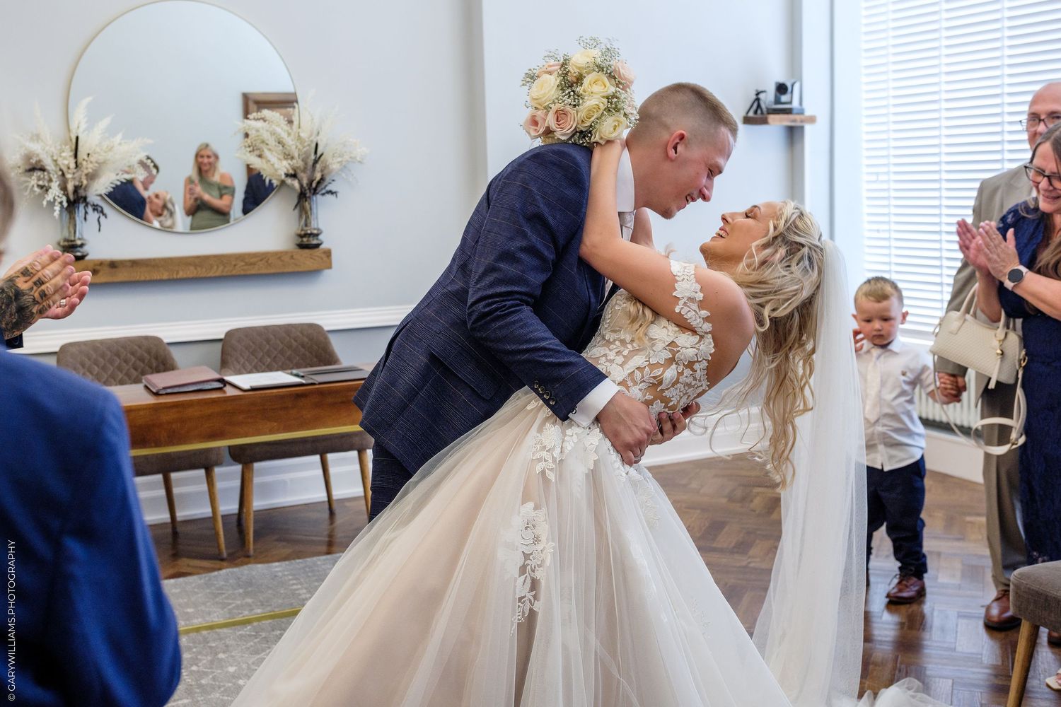 Groom dips bride for a kiss at wedding ceremony while guests applaud and a young child watches nearby.