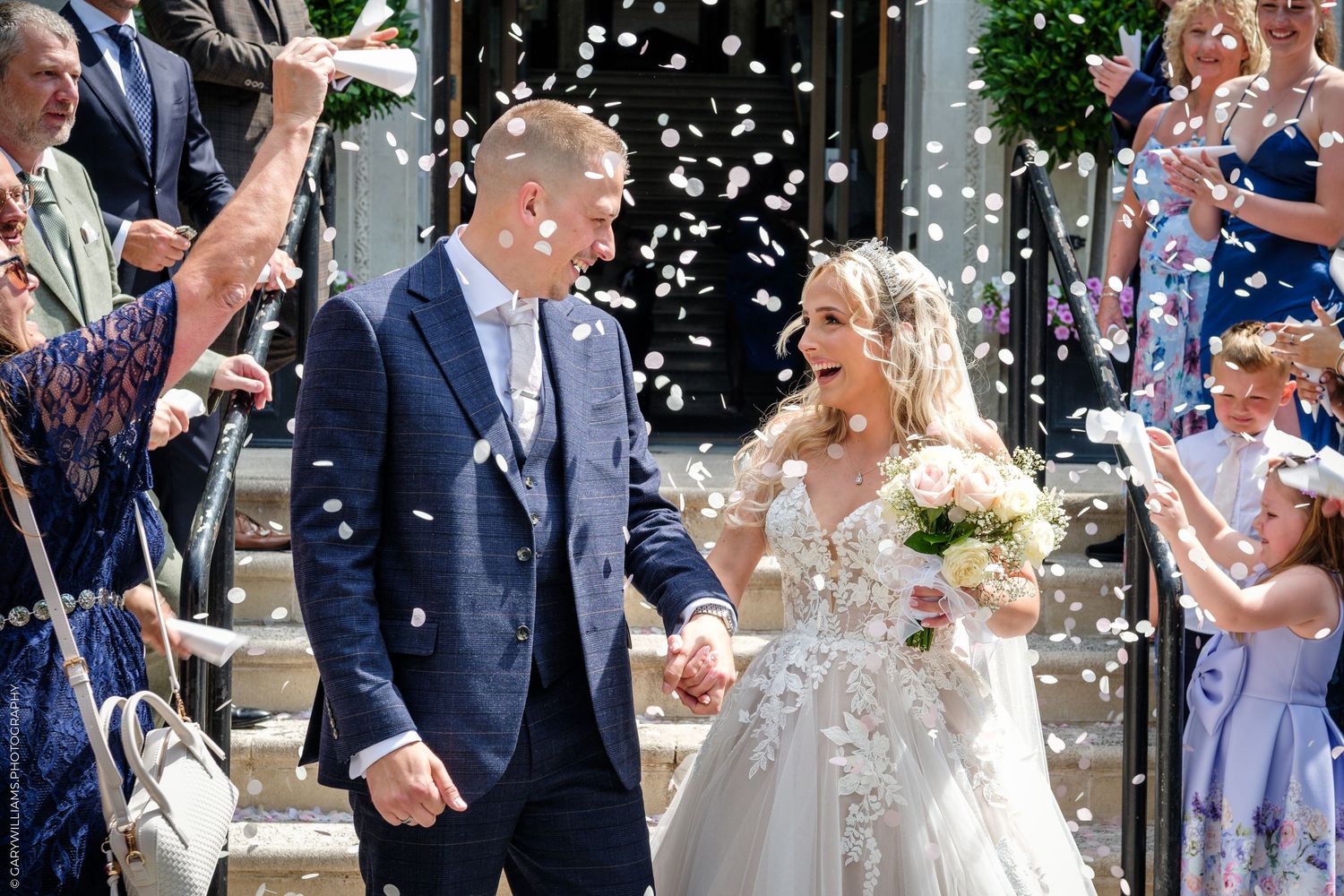 A joyful bride and groom walk through a shower of white confetti after their outdoor wedding ceremony.