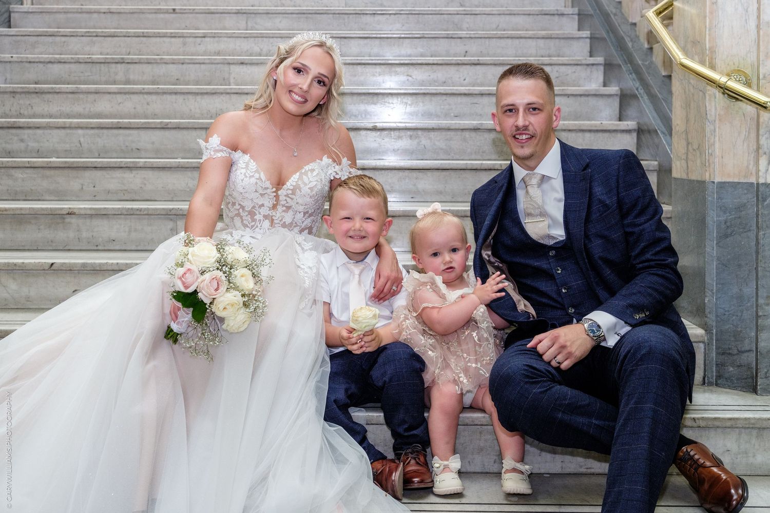 A bride and groom pose on marble stairs with two young children on their wedding day.